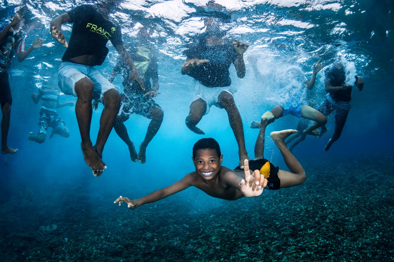 From beneath the water, we see eight boys swimming. Seven of them have their heads above water and we cannot see their faces, but one boy has swum below the water and is floating horizontally, grinning and pointing upwards.